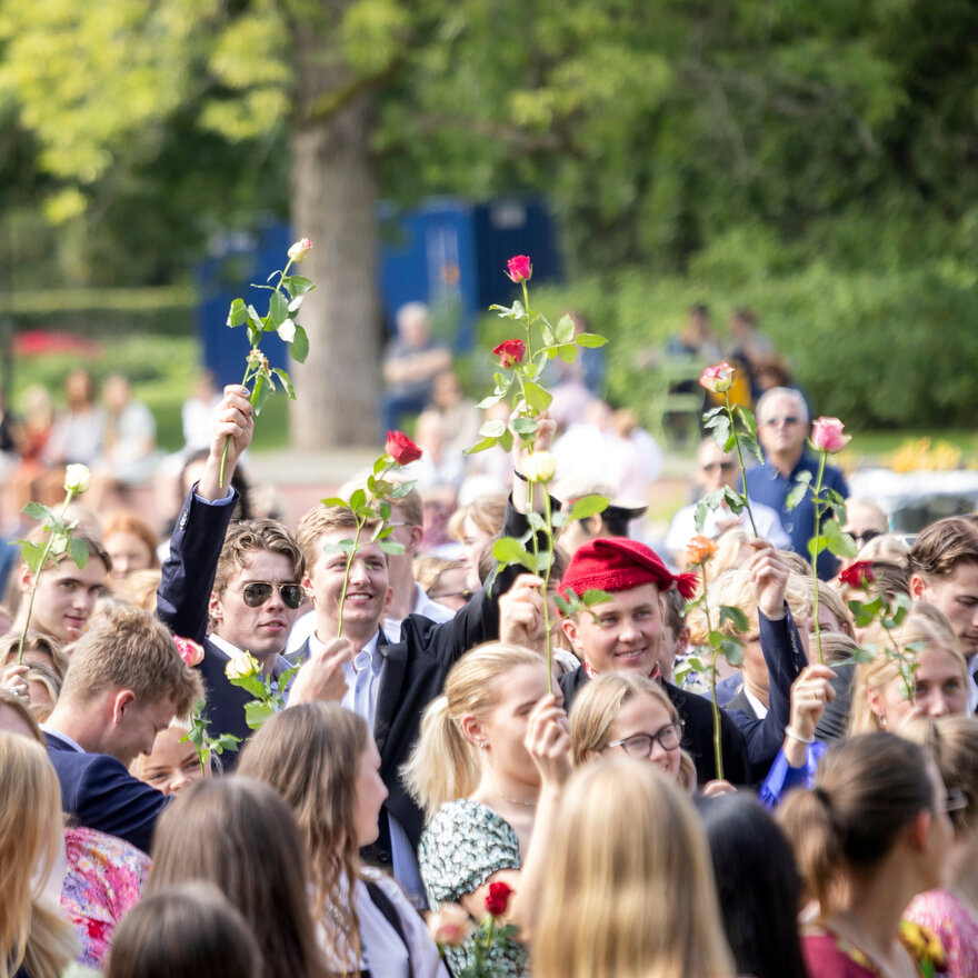 Mange studenter står ute i parken. Flere holder blomster i været. 